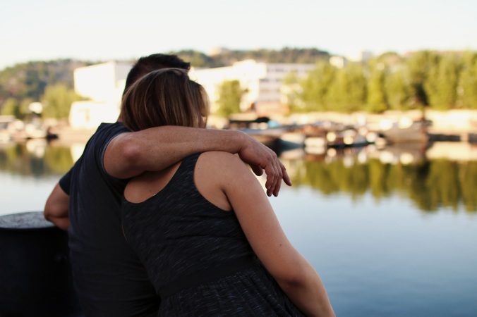 Two people sit on a bench beside the river. One puts his arm around the other while she leans her head on his shoulder.
